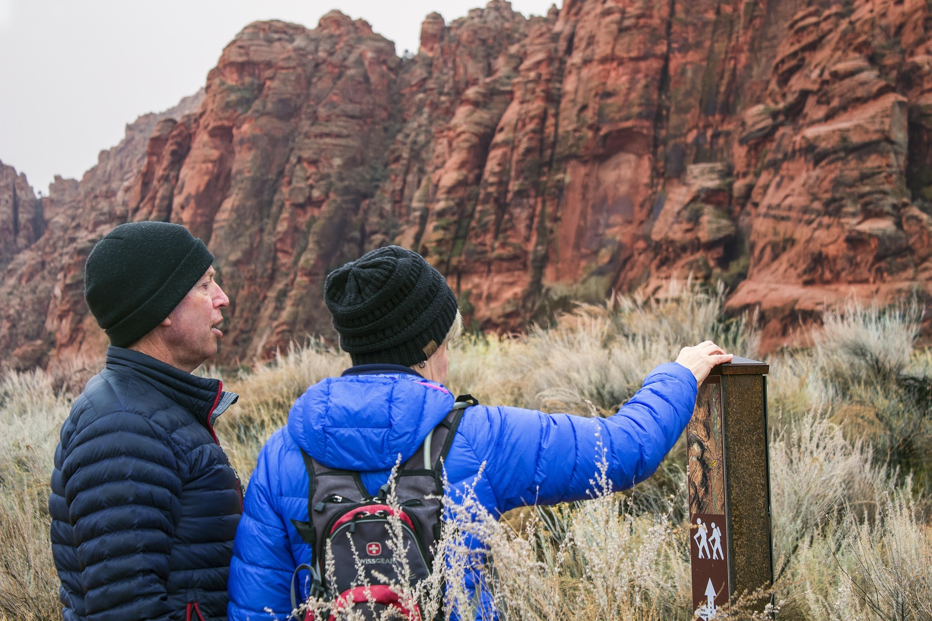 Two hikers in winter jackets read a wooden trailhead sign in front of red rock cliffs