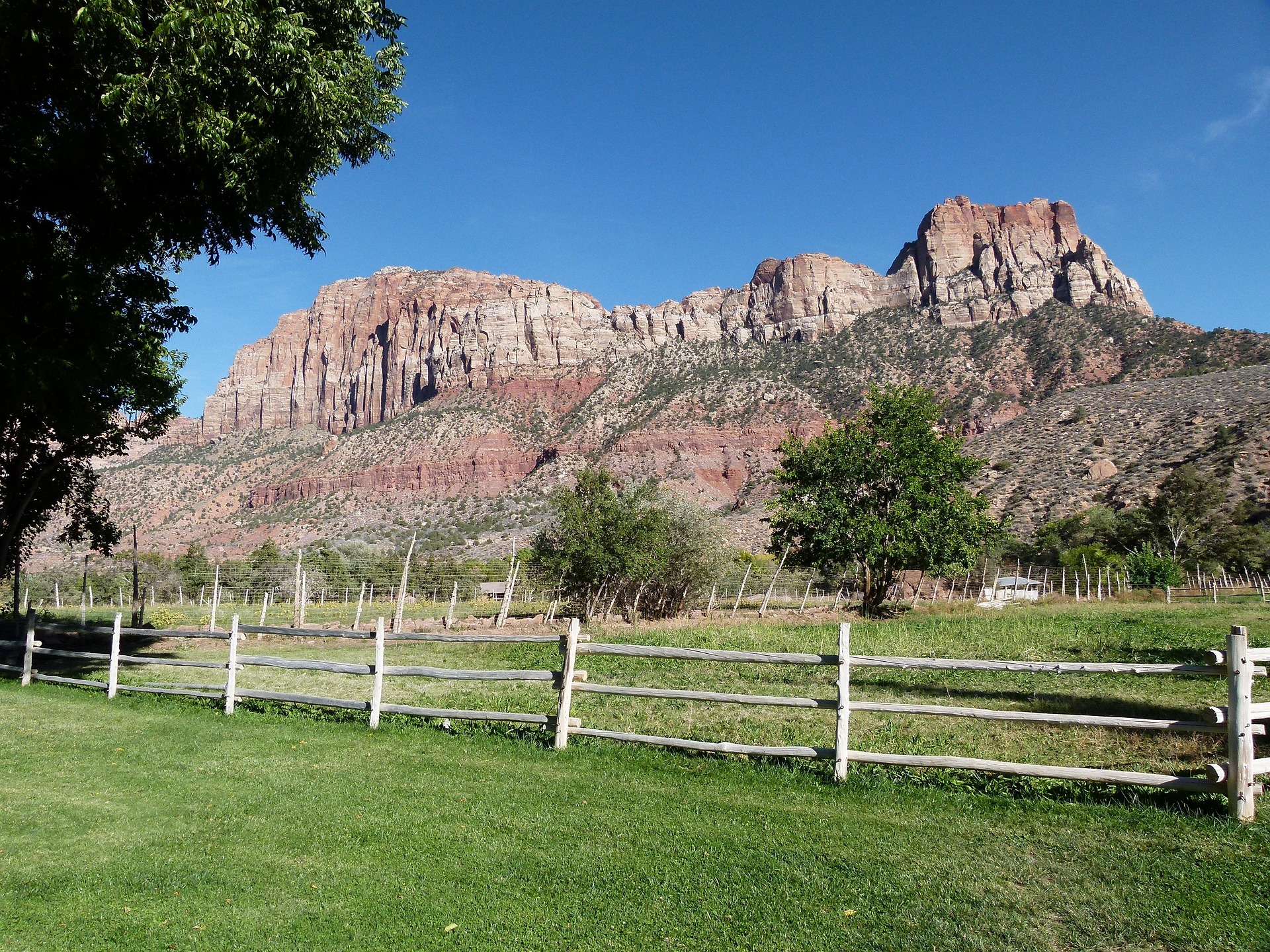 Split rail fence around a green pasture below red and white sandstone cliffs near Zion National Park