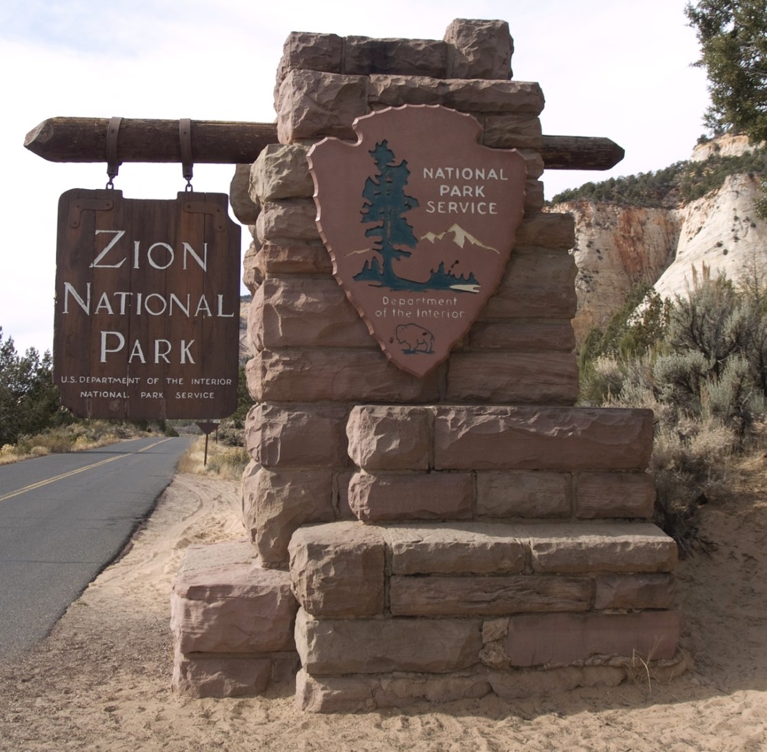 Wooden Zion National Park sign and stone National Park Service arrowhead beside the park road