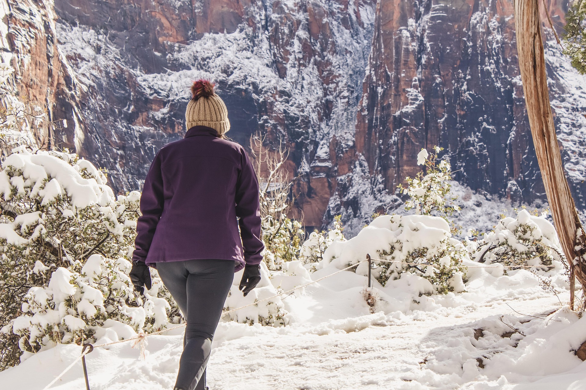 Hiker in a winter hat and fleece on a snow covered trail with a rope handrail above Zion Canyon